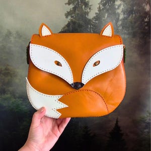 Puede incluir: Un bolso bandolera con forma de zorro hecho de cuero naranja con detalles en cuero blanco que forman la cara y la cola. El bolso presenta costuras visibles, dos orejas puntiagudas en la parte superior y una nariz de color marrón oscuro.