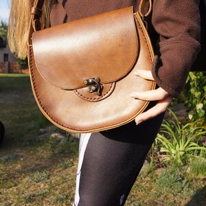 May include: A brown leather satchel bag with a flap closure and a metal buckle. The bag is worn over the shoulder of a person wearing black leggings with a skeleton pattern.
