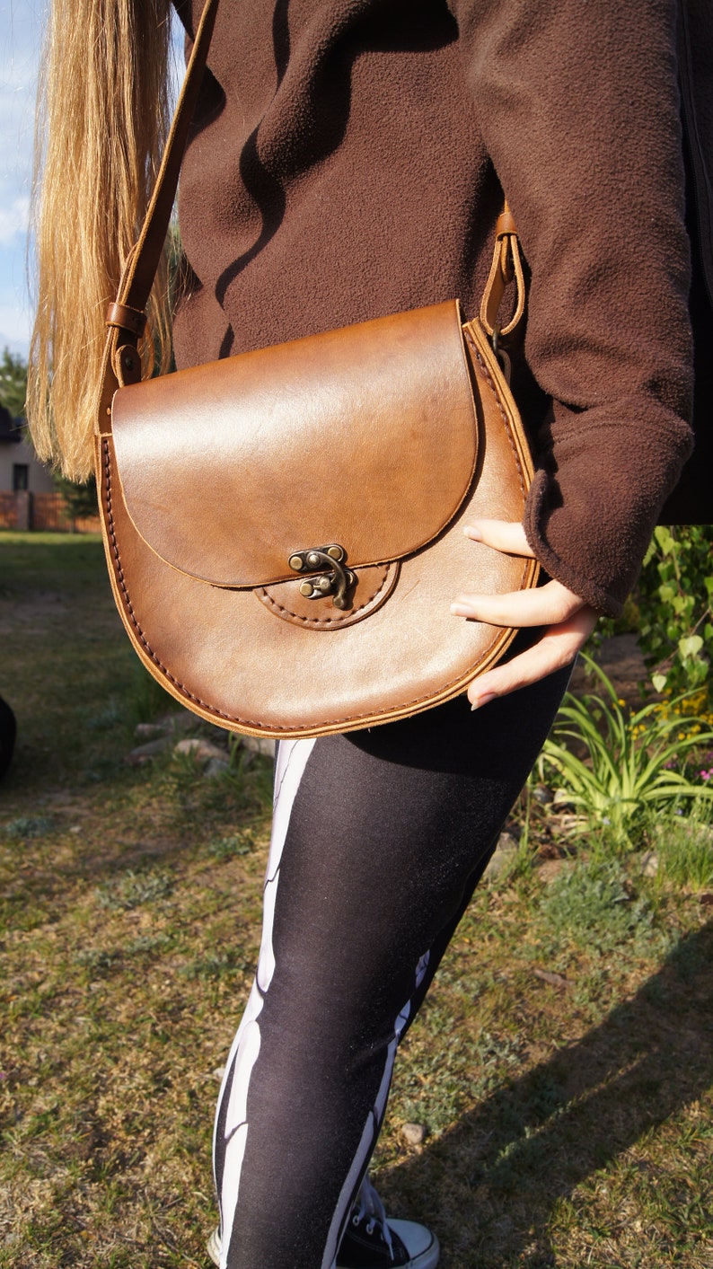 May include: A brown leather satchel bag with a flap closure and a metal buckle. The bag is worn over the shoulder of a person wearing black leggings with a skeleton pattern.