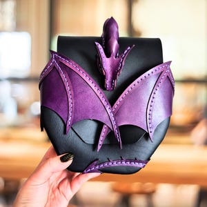 May include: A black leather pouch featuring a purple dragon design on the front. The dragon has detailed wings, a head at the top, and a tail at the bottom, all stitched onto the black base. The purple leather has a gradient finish.