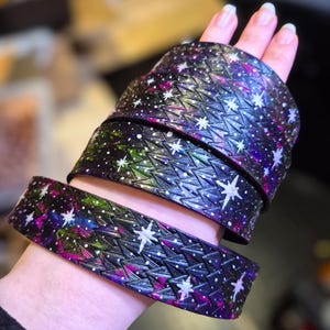 Könnte beinhalten: Ein breites Lederarmband mit einem strukturierten, schuppenartigen Muster in tiefen Schwarz-, Lila- und Grüntönen. Die Oberfläche ist mit handgemalten weißen Sternen und kleinen Punkten verziert, die einen nächtlichen Sternenhimmel darstellen.