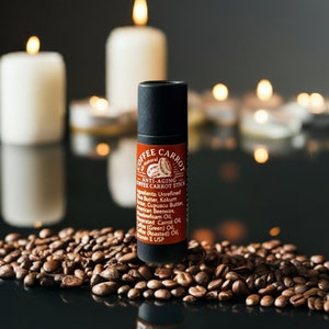 Puede incluir: Un tubo negro y rojo de "Coffee Carrot Anti-Aging Coffee Carrot Stick" descansa sobre granos de caf&eacute;. La etiqueta muestra los ingredientes. Velas encendidas en el fondo crean un ambiente c&aacute;lido. Producto para el cuidado de la piel.