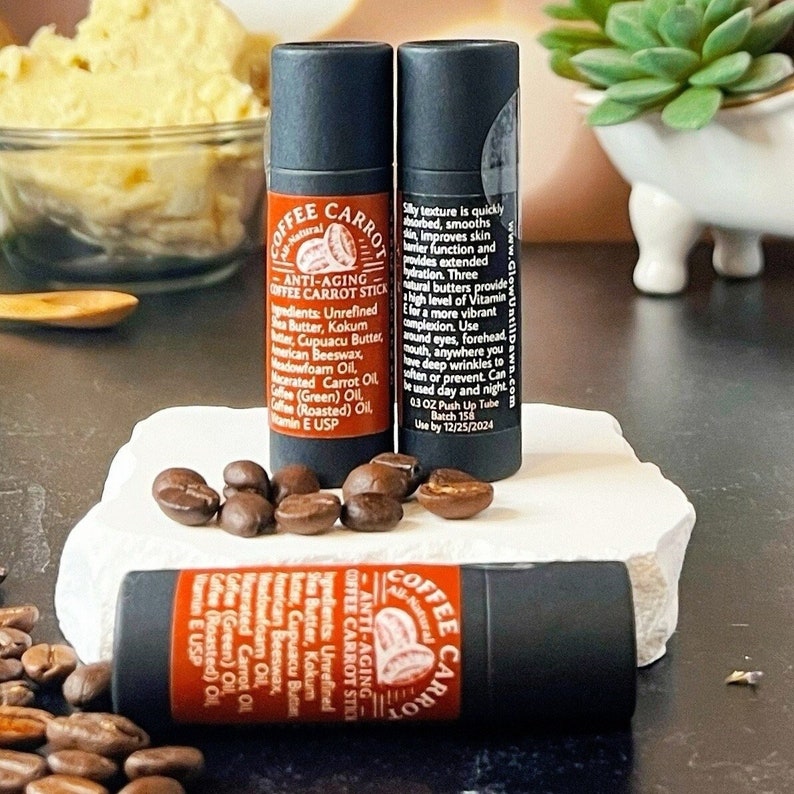 Puede incluir: Tres tubos negros de barra anti-edad "Coffee Carrot" se muestran con granos de caf&eacute;. Los tubos tienen etiquetas naranjas y negras con informaci&oacute;n del producto. La imagen tambi&eacute;n muestra un peque&ntilde;o plato de mantequilla color crema.