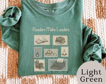 May include: A sage green long-sleeve t-shirt featuring the text "Readers Make Leaders" above a grid of six postage stamp-style illustrations depicting an open book, a stack of books, an inkwell with a quill, a coffee cup, a candle, and a typewriter.