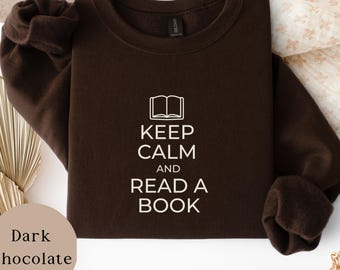 May include: A dark chocolate brown crewneck sweatshirt featuring a white graphic of an open book icon above the text "KEEP CALM AND READ A BOOK" in a clean, sans-serif font. The garment has long sleeves and a ribbed collar, cuffs, and hem.