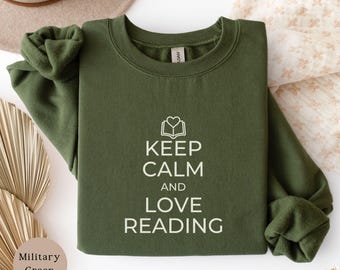 May include: A military green crewneck sweatshirt featuring a white graphic print of an open book with a heart icon above it. The text reads KEEP CALM AND LOVE READING in a clean, sans-serif font. The garment has long sleeves and a ribbed collar.