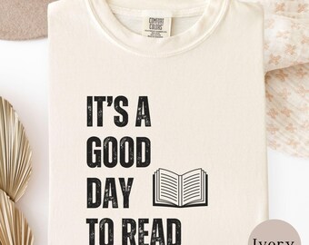 May include: An ivory crew neck t-shirt featuring the black distressed text "IT'S A GOOD DAY TO READ" stacked vertically next to a simple line drawing of an open book. The shirt has a Comfort Colors brand label printed on the inside neck area.