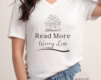 May include: A white V-neck t-shirt featuring a black line art graphic of an open book with flowers growing out of the pages. Below the illustration, the text reads Read More Worry Less in a combination of serif and script fonts with a curved line.