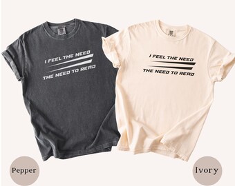 May include: Two short-sleeve crew neck t-shirts, one in pepper gray with white text and one in ivory with black text. Both shirts feature the printed phrase I FEEL THE NEED THE NEED TO READ with two horizontal lines between the two lines of text.
