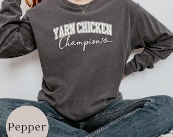 May include: A charcoal gray long-sleeve crewneck sweatshirt featuring the white text YARN CHICKEN Champion with a small heart icon. The garment has a relaxed fit and a vintage-washed appearance, paired with dark blue denim jeans.