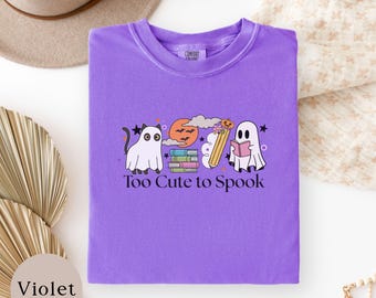 May include: A violet t-shirt featuring a graphic of a ghost cat, a stack of books, a pencil with a pumpkin, and a ghost reading a book. The text "Too Cute to Spook" is printed in black below the graphic. The shirt has a crew neckline and short sleeves.