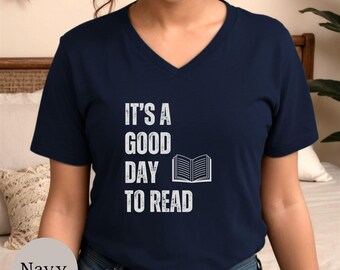 Puede incluir: Una camiseta azul marino con cuello en V que presenta texto blanco de estilo desgastado que dice IT'S A GOOD DAY TO READ, acompañado de un gráfico blanco simple de un libro abierto. La camiseta tiene mangas cortas y un corte estándar.