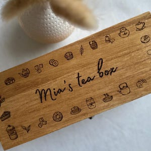 May include: A rectangular wooden tea box, featuring engraved illustrations of teacups, teapots, and pastries around the edge. The lid is inscribed with "Mia's tea box". The box is made of light brown wood, perfect for storing tea.
