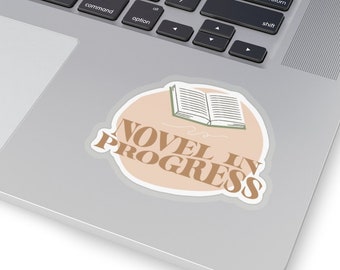 Può includere: Un adesivo con sfondo marrone chiaro e contorno bianco. L'adesivo presenta un semplice disegno a linea di un libro aperto con il testo "NOVEL IN PROGRESS" in lettere marroni.