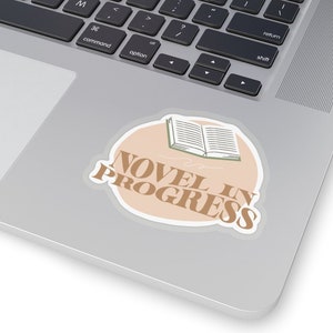 May include: A sticker with a light brown background and a white outline. The sticker features a simple line drawing of an open book with the text "NOVEL IN PROGRESS" in brown letters.