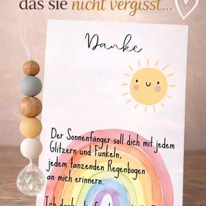 May include: A greeting card with a watercolor rainbow and a smiling sun illustration. The card has German text and is accompanied by a beaded ornament with wooden and pastel-colored beads and a crystal pendant.