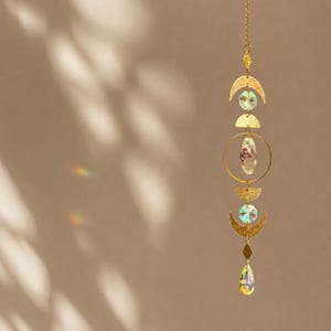 Könnte beinhalten: Ein goldfarbener Sonnenfänger mit einem himmlischen Design. Er verfügt über Halbmonde, einen Kreis und einen tropfenförmigen Kristall, die alle durch eine zarte Kette verbunden sind. Der Sonnenfänger enthält auch klare, facettierte Kristalle und ein zentrales Oval mit getrockneten Blumen.