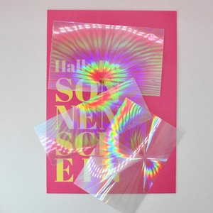 Könnte beinhalten: Ein rosa Poster mit dem Wort "SONNENSCHEIN" in Gelb, überlagert mit schillernden, transparenten Folien. Die Folien erzeugen einen Regenbogeneffekt mit einem radialen Design oben. Das Wort "HALLO" ist oben sichtbar.