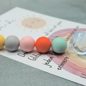 May include: A card with a watercolor rainbow, sun, and text, accompanied by colorful round beads and a faceted crystal sphere. The beads are yellow, gray, pink, orange, and mint green. The card has German text.