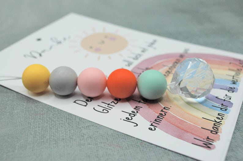 May include: A card with a watercolor rainbow, sun, and text, accompanied by colorful round beads and a faceted crystal sphere. The beads are yellow, gray, pink, orange, and mint green. The card has German text.
