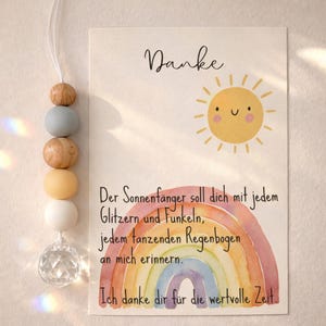 May include: A decorative suncatcher with wooden and pastel-colored beads, and a clear crystal. A card with a watercolor rainbow, a smiling sun, and the German text "Danke" and a message.