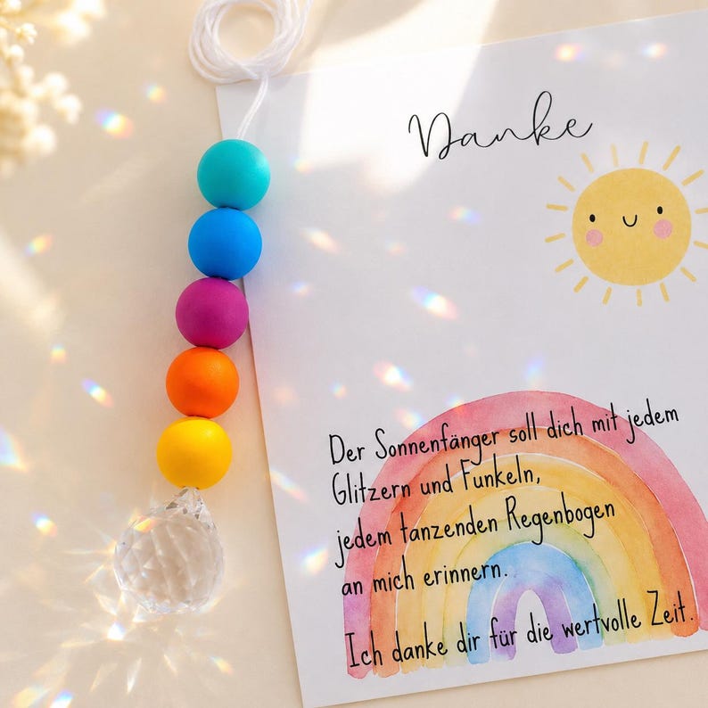 May include: A suncatcher featuring a string of colorful beads in teal, blue, purple, orange, and yellow, ending in a clear faceted crystal prism. Beside it, a white card displays a yellow sun illustration and German text with a watercolor rainbow.