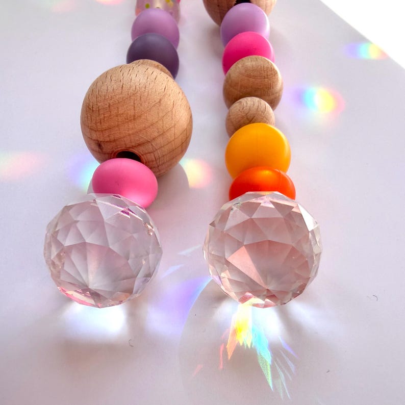 May include: Two strands of decorative beads in a rainbow of colors. The strands include wooden, pink, purple, orange, and clear crystal beads. The beads are arranged on a white surface, with light refracting through the crystal beads.