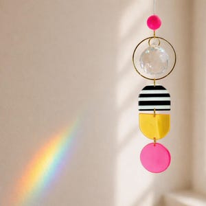 Könnte beinhalten: Ein hängender geometrischer Sonnenfänger mit einer durchsichtigen Kristallkugel, goldfarbenen Ringen und farbigen Acrylformen. Das Design umfasst einen schwarz-weiß gestreiften Halbkreis, einen gelben Halbkreis und einen rosa Kreis. Ein Regenbogenprisma wird an die Wand geworfen.