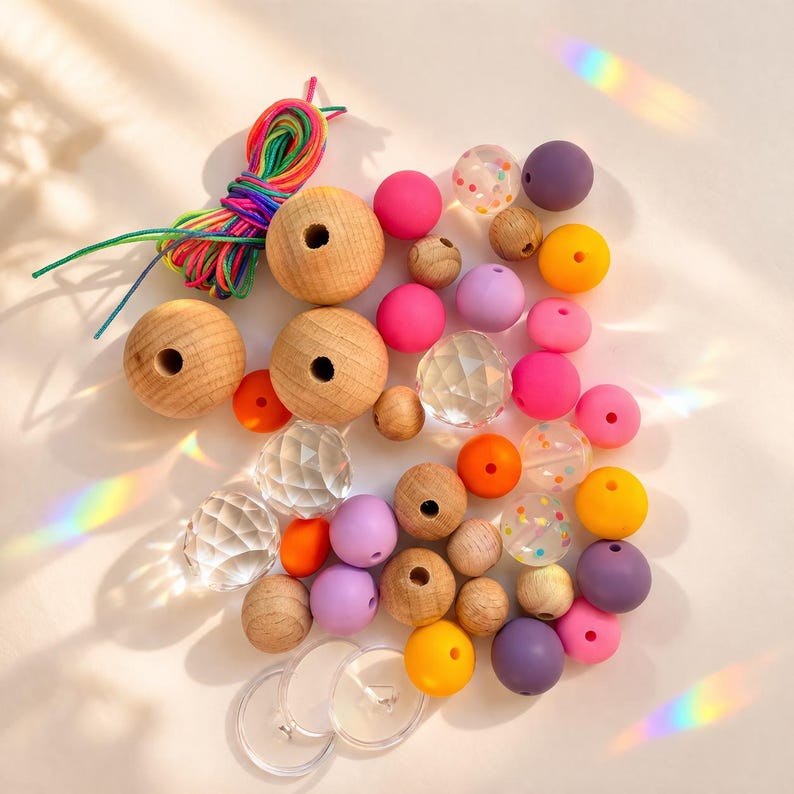 May include: A collection of craft beads in various colors and materials. Includes large wooden beads, pink, purple, and yellow silicone beads, clear faceted beads, and small wooden beads. A bundle of rainbow-colored string is also present, along with clear plastic discs.