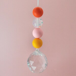 May include: A crystal suncatcher with a string of colorful beads. The beads are orange, pink, clear with confetti, and yellow.