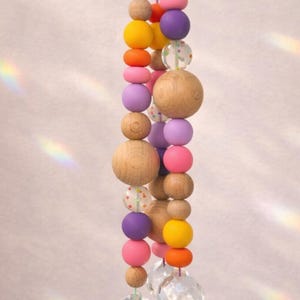 May include: A hanging decorative mobile featuring a string of wooden beads in various sizes and colors, including natural wood, pink, purple, orange, and yellow. Clear crystal prisms hang at the bottom, reflecting light.