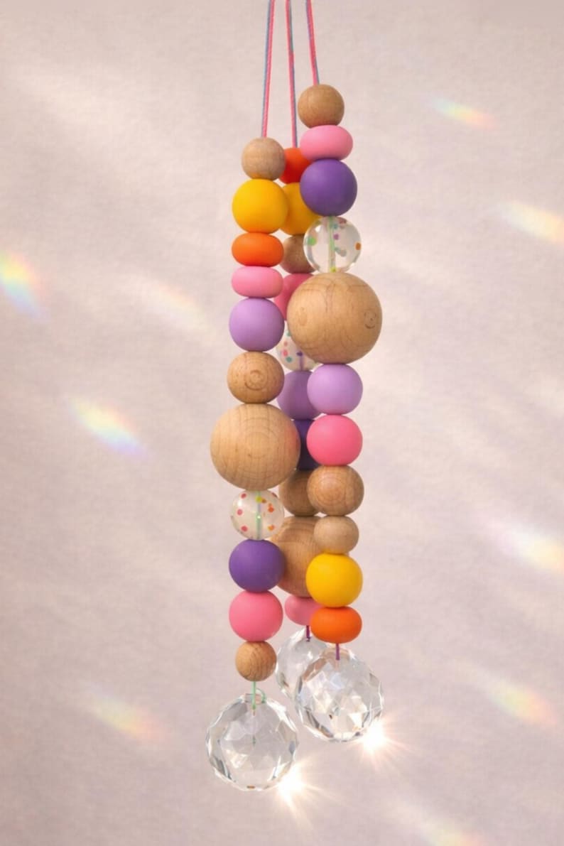 May include: A hanging decorative mobile featuring a string of wooden beads in various sizes and colors, including natural wood, pink, purple, orange, and yellow. Clear crystal prisms hang at the bottom, reflecting light.