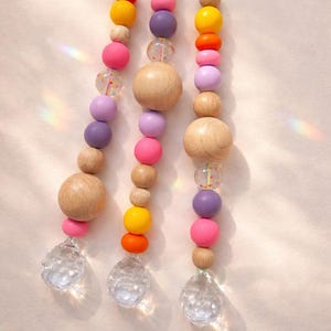 May include: Three strands of colorful wooden beads in shades of pink, purple, orange, and yellow, interspersed with clear, faceted crystal pendants. The beads are strung on colorful cords, creating a playful and decorative item.