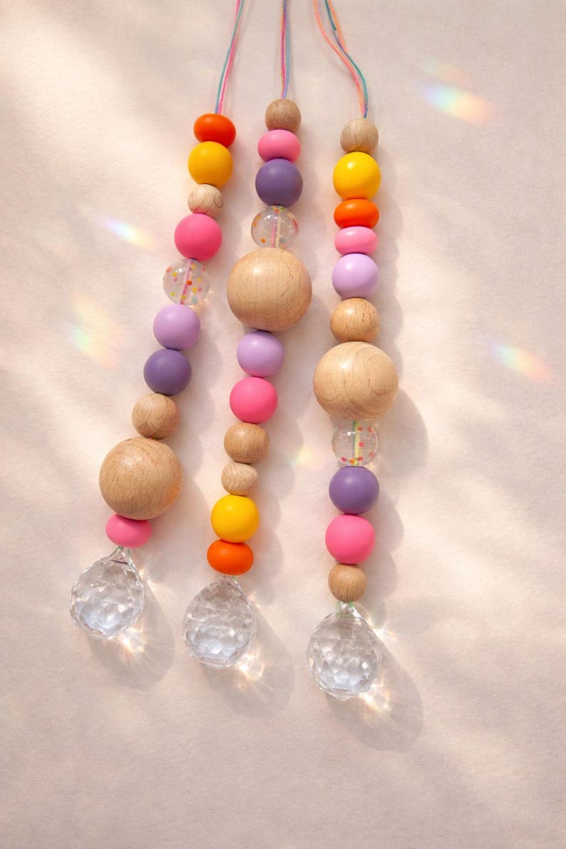 May include: Three strands of colorful wooden beads in shades of pink, purple, orange, and yellow, interspersed with clear, faceted crystal pendants. The beads are strung on colorful cords, creating a playful and decorative item.