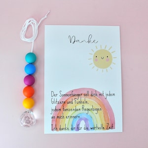 May include: A suncatcher featuring a white string with six colorful beads in teal, blue, purple, orange, and yellow, ending in a clear faceted crystal. Beside it is a white card with a yellow sun illustration and a watercolor rainbow.