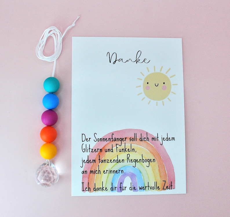 May include: A suncatcher featuring a white string with six colorful beads in teal, blue, purple, orange, and yellow, ending in a clear faceted crystal. Beside it is a white card with a yellow sun illustration and a watercolor rainbow.