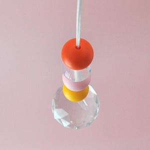 May include: A crystal prism hanging from a white cord with three colorful beads. The beads are orange, pink with blue polka dots, and yellow.