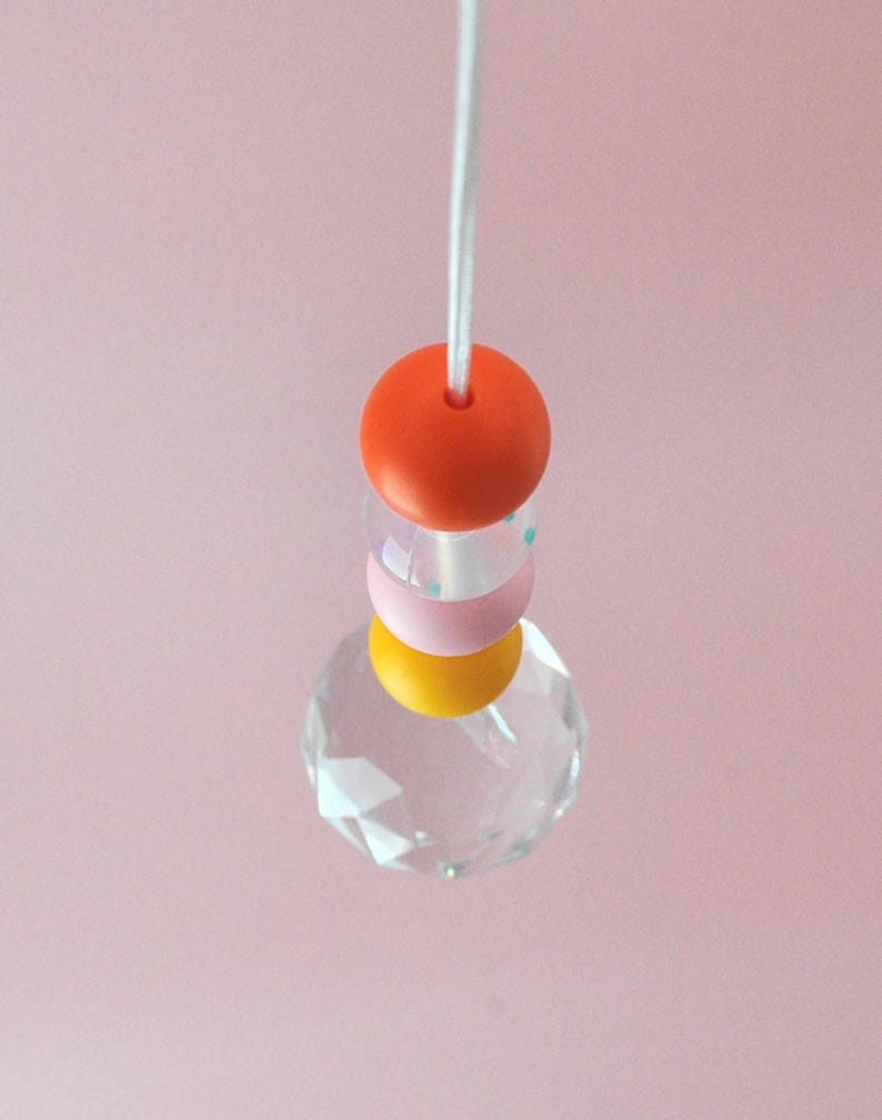 May include: A crystal prism hanging from a white cord with three colorful beads. The beads are orange, pink with blue polka dots, and yellow.