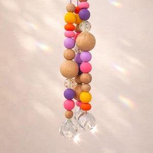 May include: A hanging decorative mobile featuring a string of colorful wooden beads in shades of pink, purple, yellow, and orange, interspersed with clear crystal pendants. The beads vary in size and shape.