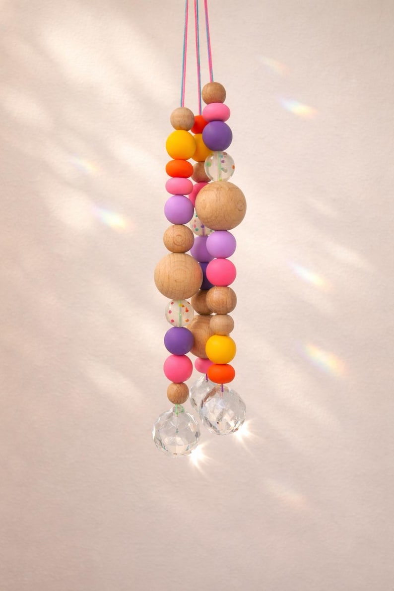 May include: A hanging decorative mobile featuring a string of colorful wooden beads in shades of pink, purple, yellow, and orange, interspersed with clear crystal pendants. The beads vary in size and shape.
