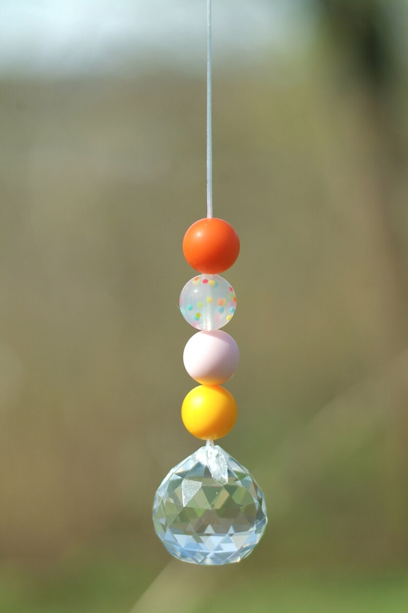May include: A crystal suncatcher with a string of colorful beads. The beads are orange, white with colorful dots, pink, and yellow.