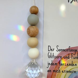 May include: A hanging suncatcher with a clear crystal ball at the bottom and wooden and white beads. The suncatcher is suspended by a white string. A card with German text and a rainbow illustration is in the background.