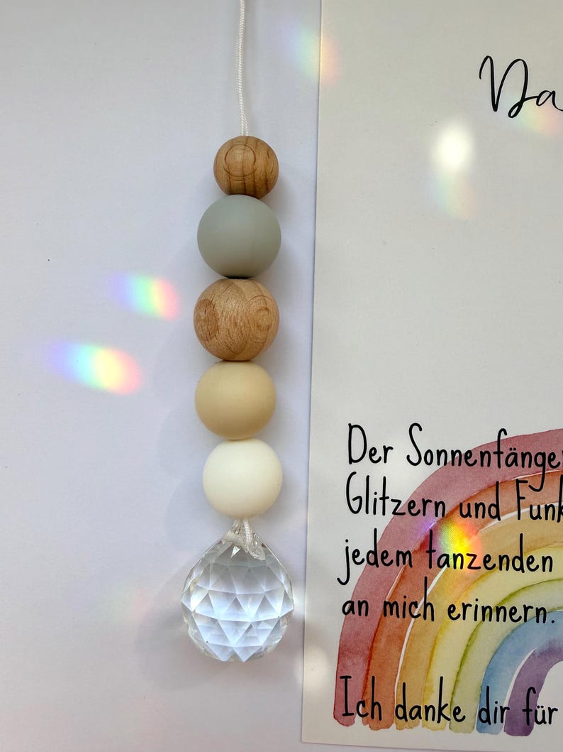 May include: A hanging suncatcher with a clear crystal ball at the bottom and wooden and white beads. The suncatcher is suspended by a white string. A card with German text and a rainbow illustration is in the background.
