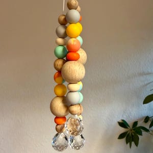 May include: A hanging decorative mobile featuring a string of wooden beads in various sizes and colors, including natural wood, gray, yellow, orange, and mint green. Two clear crystal spheres hang at the bottom.