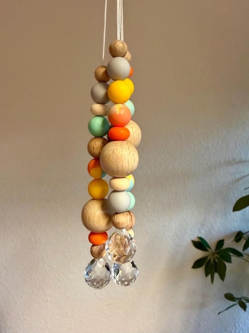 May include: A hanging decorative mobile featuring a string of wooden beads in various sizes and colors, including natural wood, gray, yellow, orange, and mint green. Two clear crystal spheres hang at the bottom.