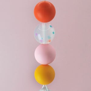 May include: A silicone bead necklace with four beads. The top bead is orange, the second bead is clear with colorful confetti inside, the third bead is pink, and the bottom bead is yellow.