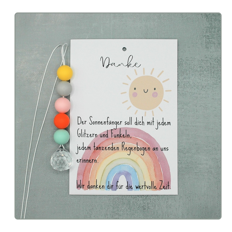 May include: A decorative card with the word "Danke" and a smiling sun illustration. The card features a rainbow and text in German. A string of colorful beads and a crystal ball hang from the card.