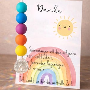 May include: A suncatcher featuring a string of five colorful silicone beads in turquoise, blue, purple, orange, and yellow, ending in a clear faceted crystal prism. Beside it stands a white card with a sun illustration and German text.