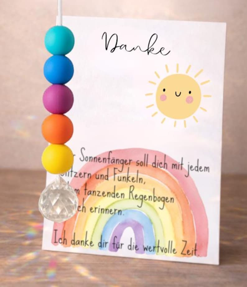 May include: A suncatcher featuring a string of five colorful silicone beads in turquoise, blue, purple, orange, and yellow, ending in a clear faceted crystal prism. Beside it stands a white card with a sun illustration and German text.