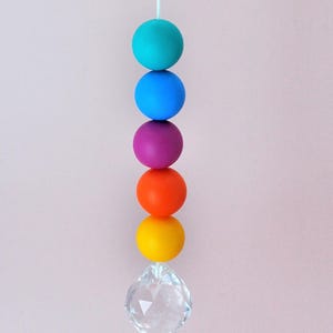 May include: A vertical hanging ornament featuring five matte spherical beads in turquoise, blue, purple, orange, and yellow, suspended from a white string. A clear, multifaceted crystal prism hangs at the bottom of the colorful bead stack.